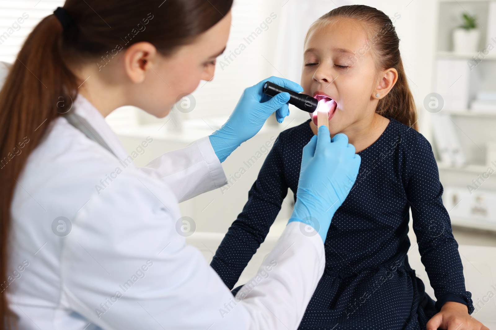 Photo of Doctor examining girl's throat with flashlight and tongue depressor in clinic