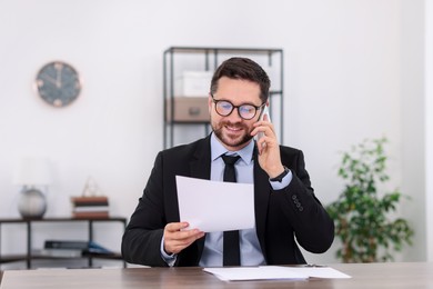 Photo of Banker with document talking on smartphone at wooden table in office