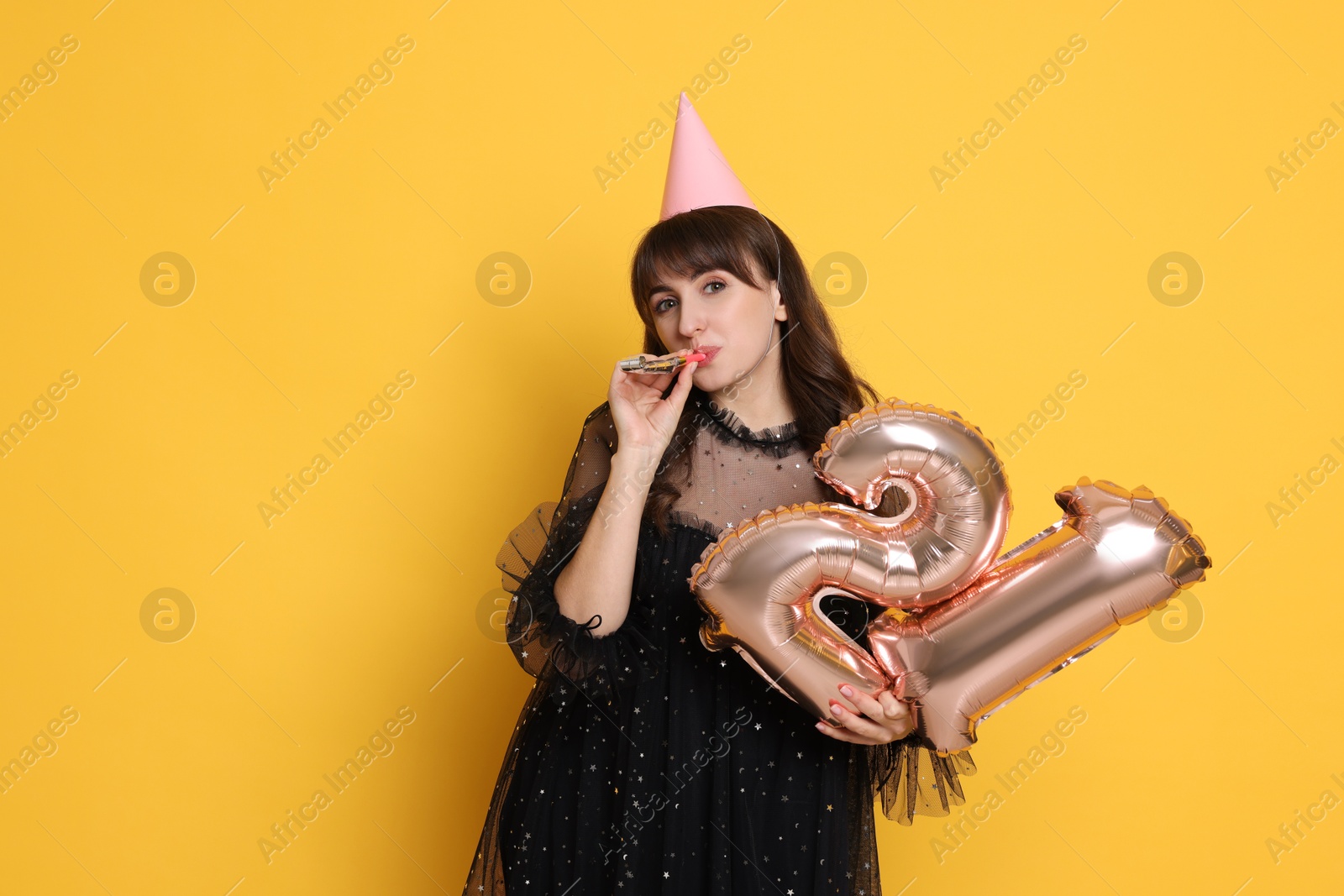 Photo of Coming of age party - 21st birthday. Young woman with hat and blower holding number shaped balloons on yellow background