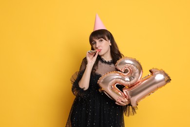 Photo of Coming of age party - 21st birthday. Young woman with hat and blower holding number shaped balloons on yellow background