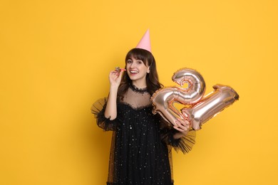 Coming of age party - 21st birthday. Young woman with hat and blower holding number shaped balloons on yellow background