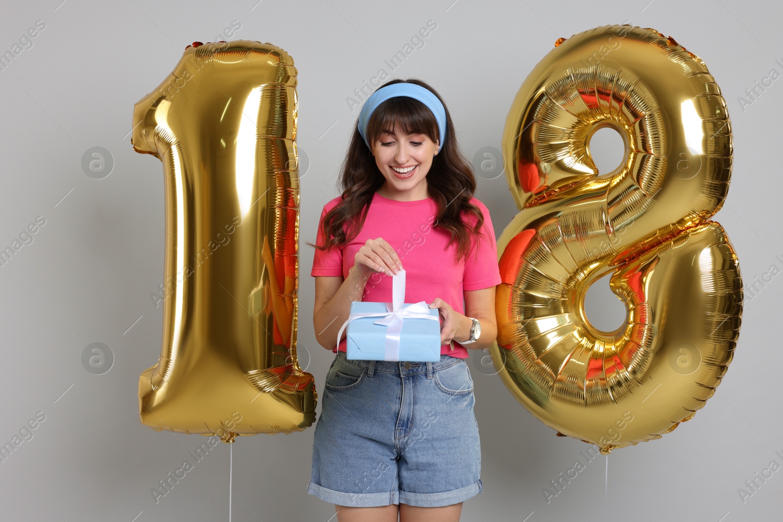 Photo of Coming of age party - 18th birthday. Young woman with gift box holding number shaped balloons on light grey background