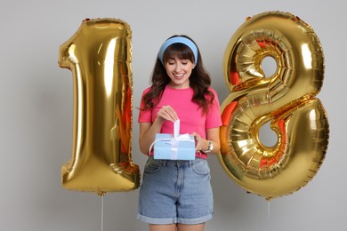 Photo of Coming of age party - 18th birthday. Young woman with gift box holding number shaped balloons on light grey background