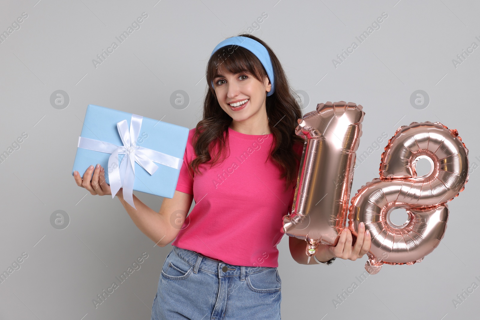 Photo of Coming of age party - 18th birthday. Young woman with gift box holding number shaped balloons on light grey background