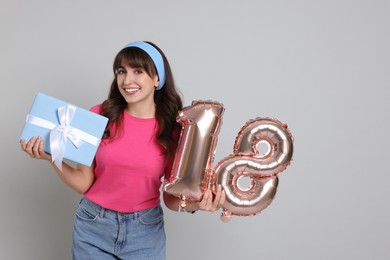 Photo of Coming of age party - 18th birthday. Young woman with gift box holding number shaped balloons on light grey background, space for text