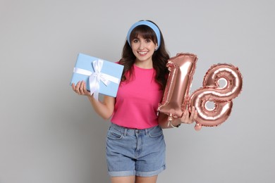 Coming of age party - 18th birthday. Young woman with gift box holding number shaped balloons on light grey background