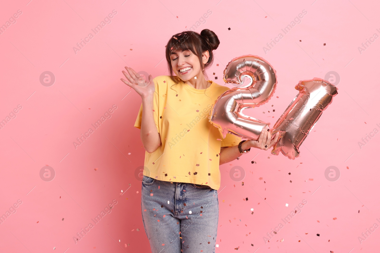 Photo of Coming of age party - 21st birthday. Young woman holding number shaped balloons on pink background with confetti