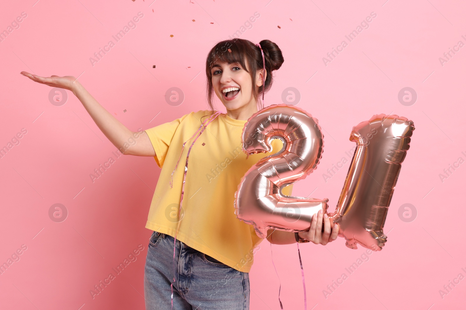 Photo of Coming of age party - 21st birthday. Young woman holding number shaped balloons on pink background with confetti