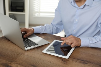 Photo of Businessman using laptop and tablet at wooden table, closeup. Modern technology