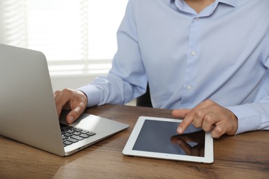 Photo of Businessman using laptop and tablet at wooden table, closeup. Modern technology