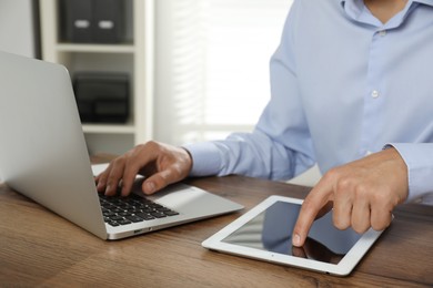 Photo of Businessman using laptop and tablet at wooden table, closeup. Modern technology