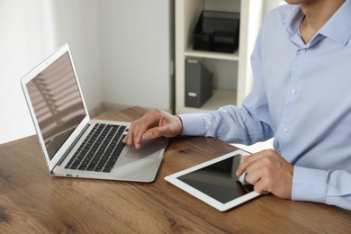 Photo of Businessman using laptop and tablet at wooden table, closeup. Modern technology