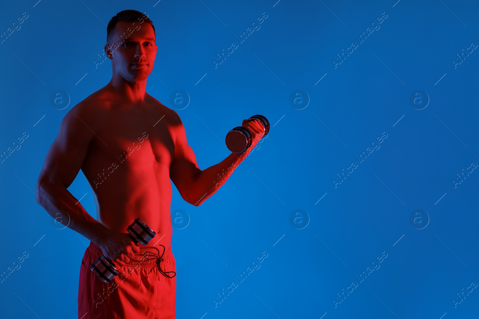 Photo of Man exercising with dumbbells in red light on blue background, space for text