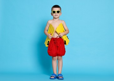 Cute little boy in beachwear with sunglasses and fins on light blue background