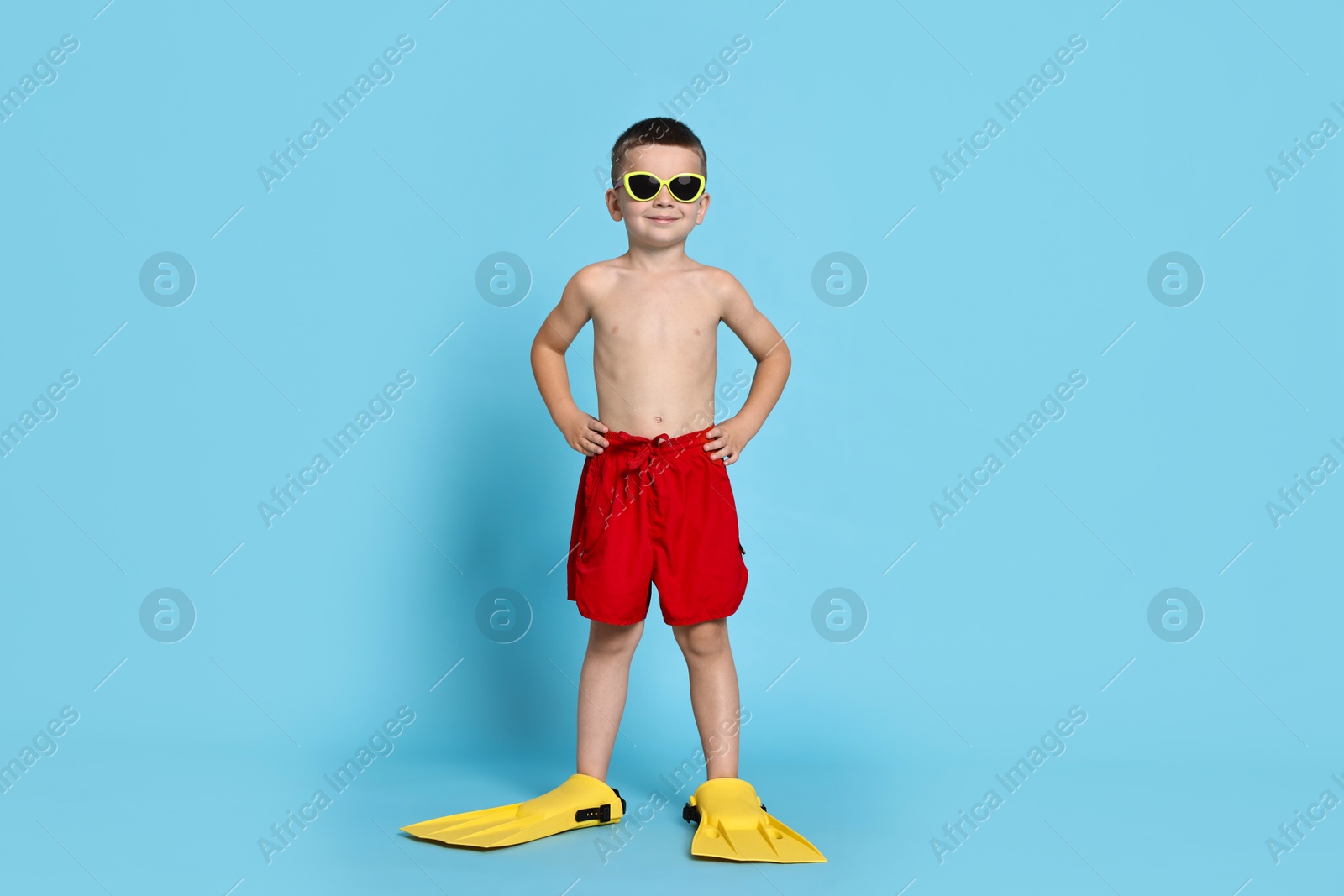 Photo of Cute little boy in beachwear with sunglasses and fins on light blue background