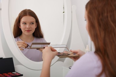 Photo of Teenage girl with eyeshadow palette near mirror indoors