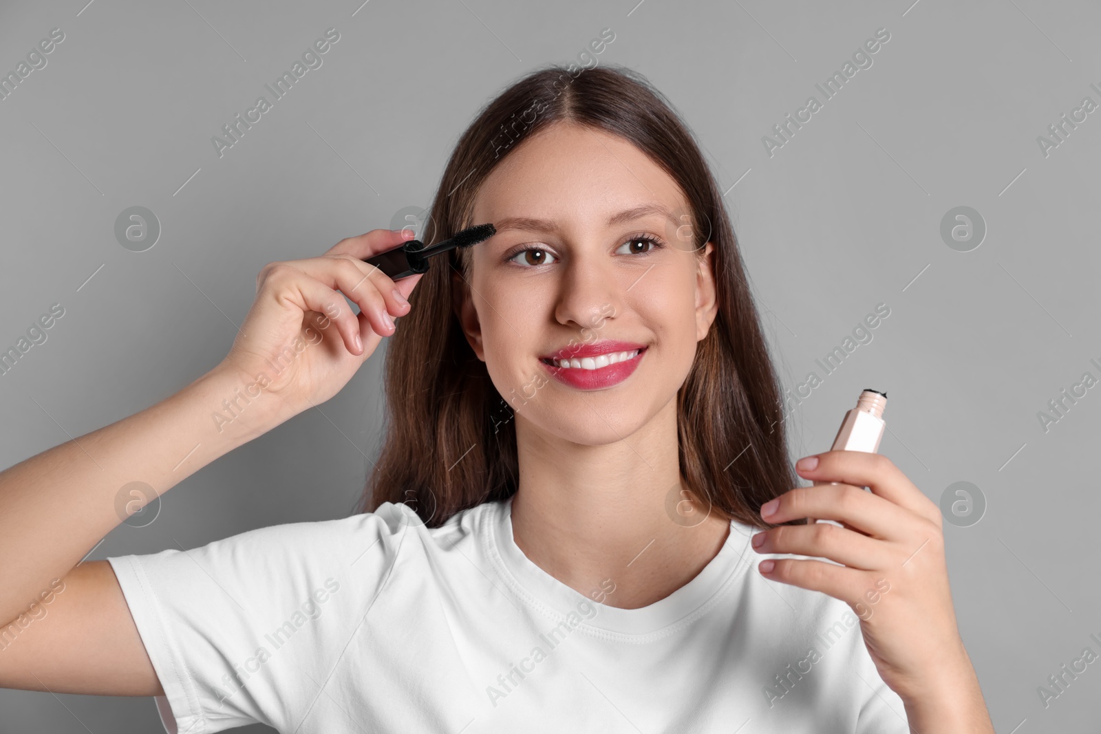 Photo of Smiling teenage girl applying mascara on grey background