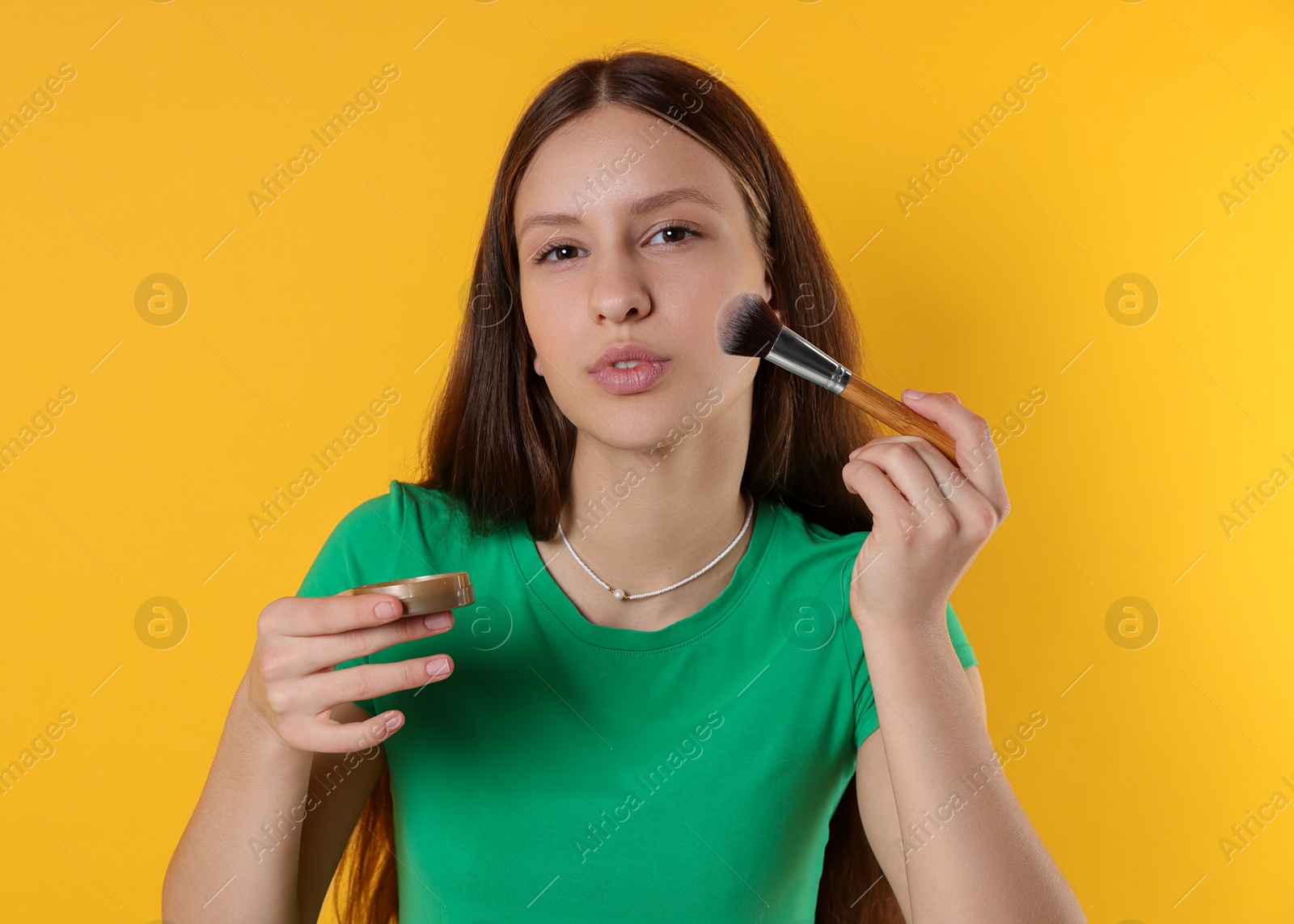 Photo of Teenage girl applying blusher with makeup brush on yellow background