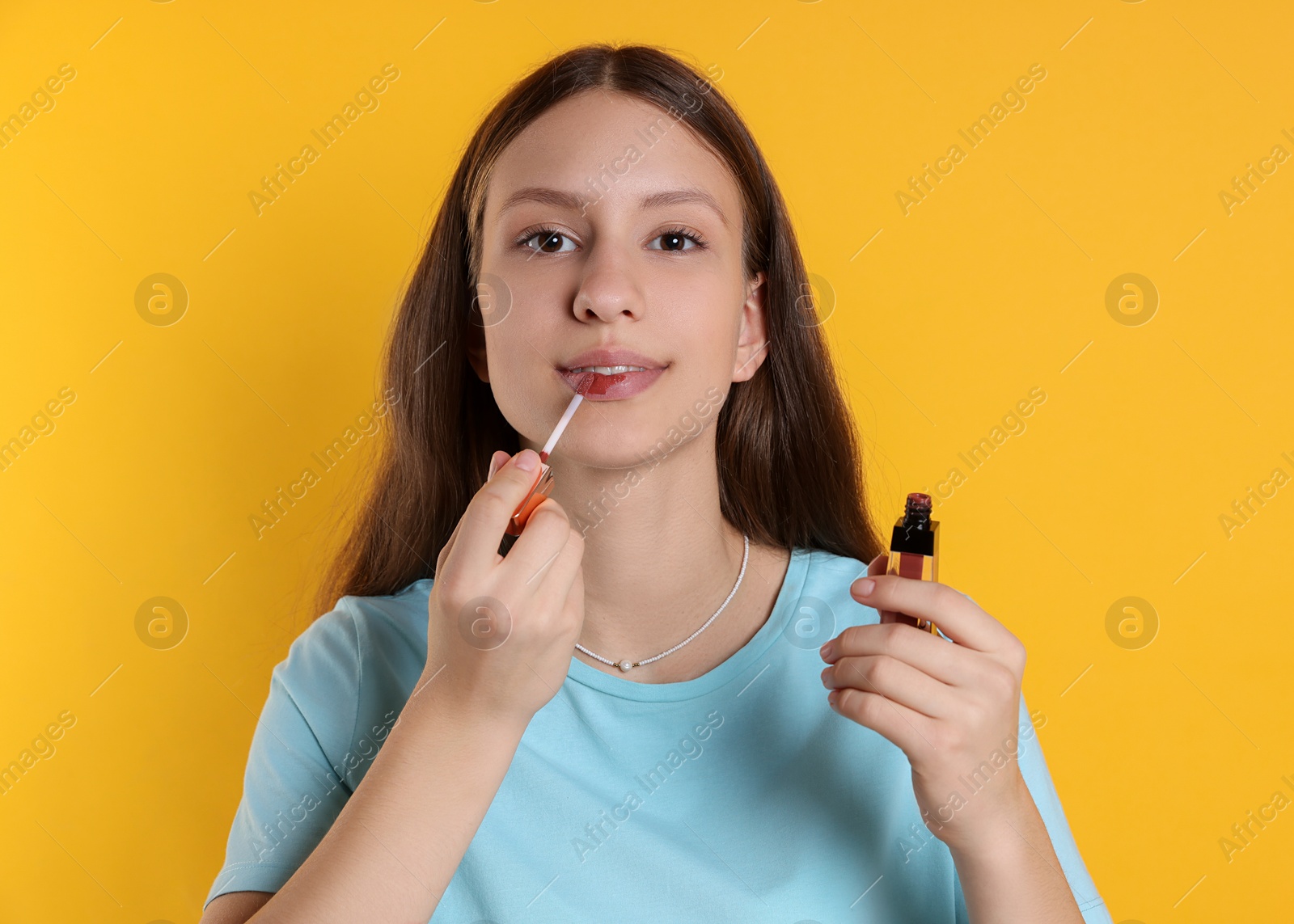 Photo of Teenage girl applying lip gloss on yellow background