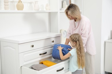 Photo of Little girl helping her mother putting clothes into drawers at home