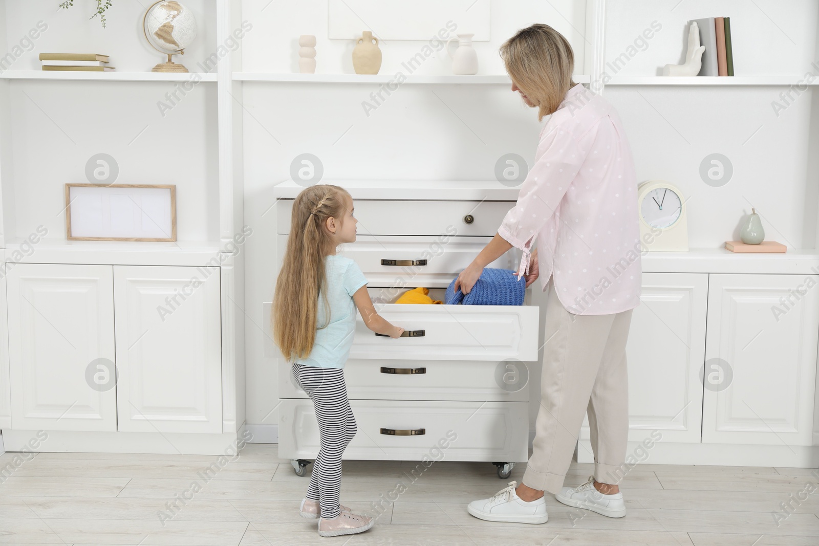 Photo of Little girl helping her mother putting clothes into drawers at home