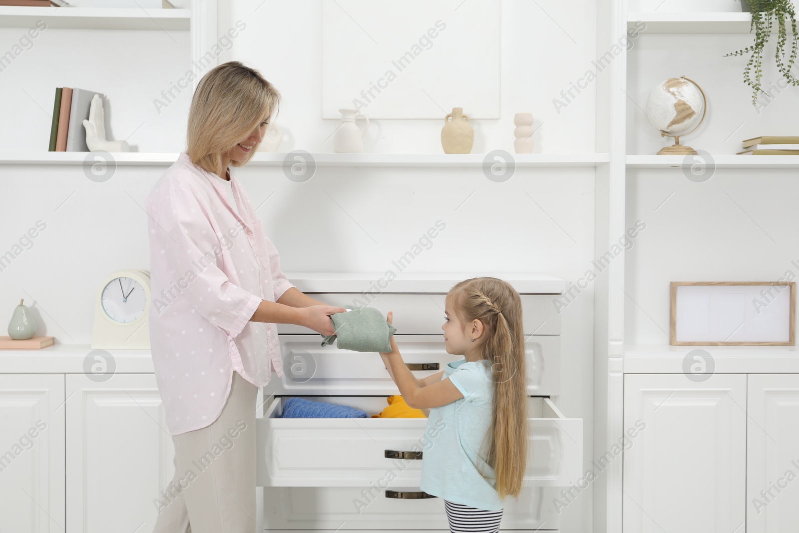 Photo of Little girl helping her mother putting clothes into drawers at home