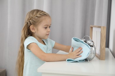 Photo of Little helper. Cute girl wiping dust from alarm clock at home