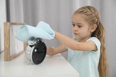 Photo of Little helper. Cute girl wiping dust from alarm clock at home