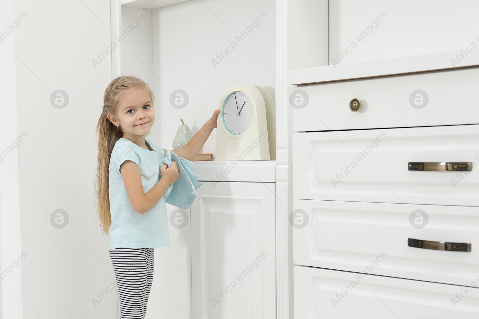 Photo of Little helper. Cute girl wiping dust from clock at home