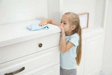 Photo of Little helper. Cute girl wiping dust from chest of drawers at home