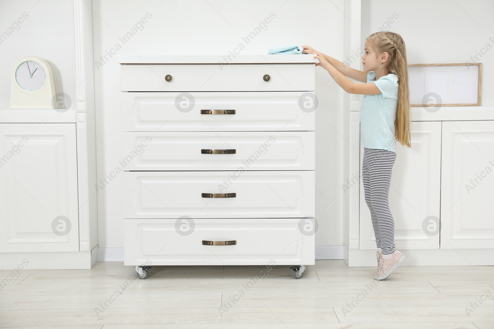 Photo of Little helper. Cute girl wiping dust from chest of drawers at home