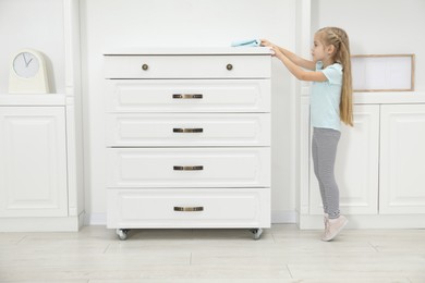 Photo of Little helper. Cute girl wiping dust from chest of drawers at home