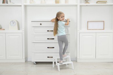 Photo of Little helper. Cute girl wiping dust from chest of drawers at home