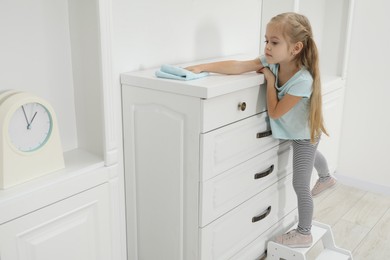 Photo of Little helper. Cute girl wiping dust from chest of drawers at home