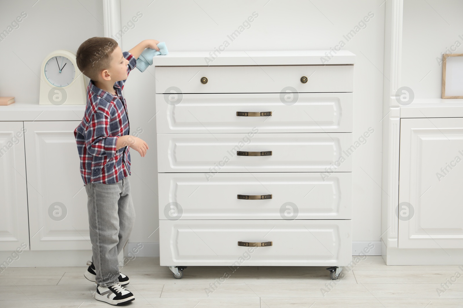 Photo of Little helper. Cute boy wiping dust from chest of drawers at home