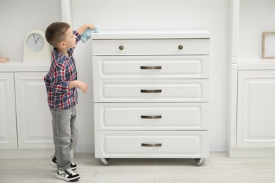 Photo of Little helper. Cute boy wiping dust from chest of drawers at home