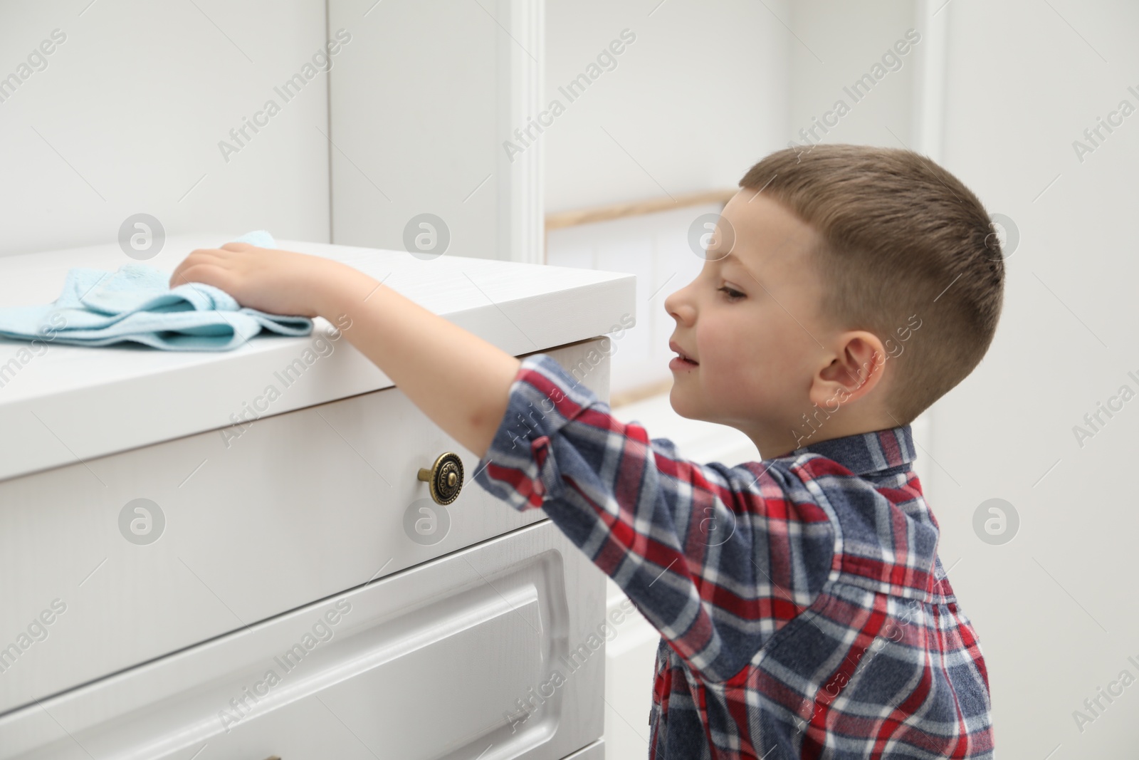 Photo of Little helper. Cute boy wiping dust from chest of drawers at home