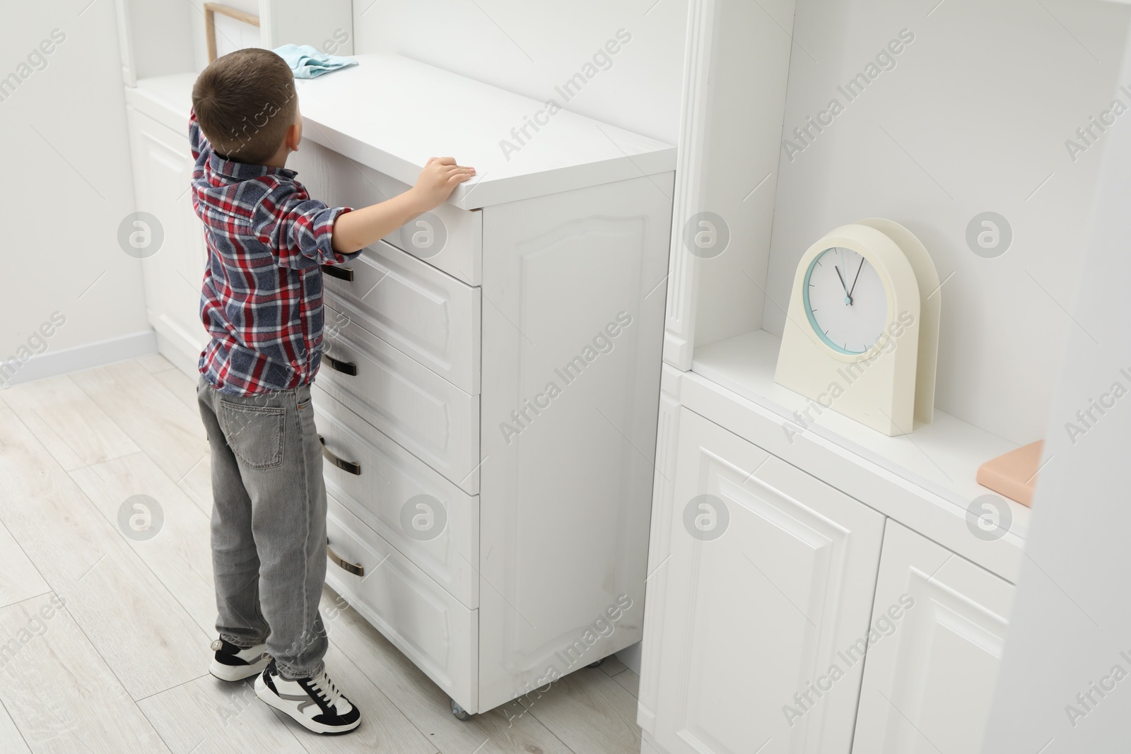 Photo of Little helper. Cute boy wiping dust from chest of drawers at home, back view