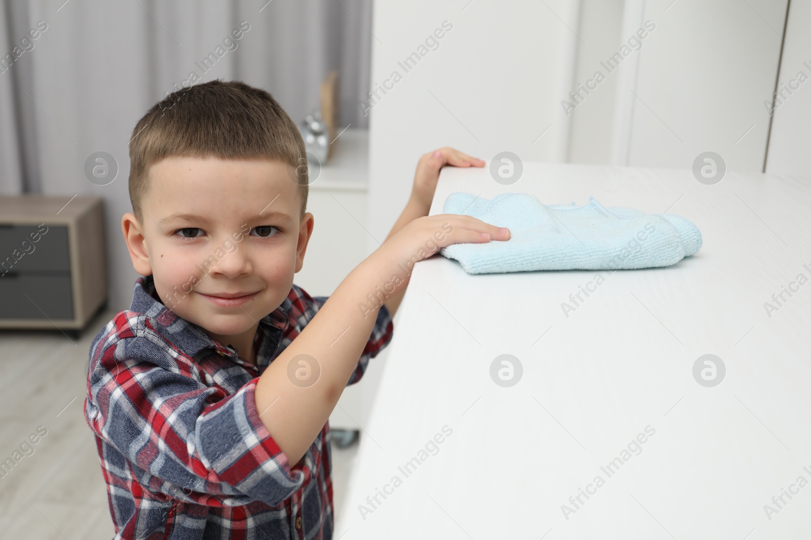 Photo of Little helper. Cute boy wiping dust from chest of drawers at home