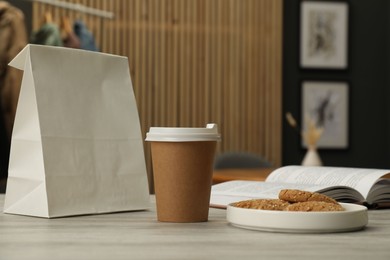 Photo of Paper cup, plate of cookies, book and takeaway bag on light wooden table in cafe. Mockup for design