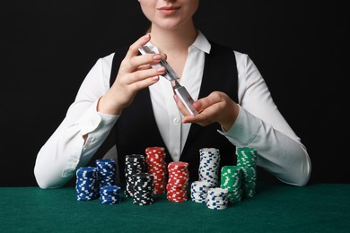 Professional croupier with chips shuffling playing cards at gambling table on black background, closeup