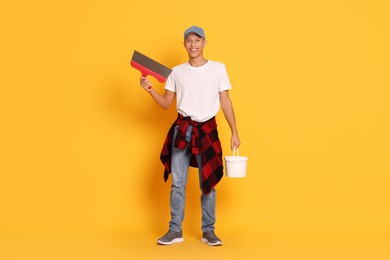 Young decorator with putty knife and bucket on orange background