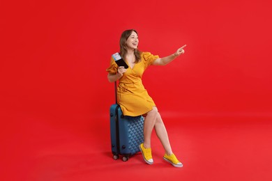 Happy traveller with passport and ticket sitting on suitcase against red background