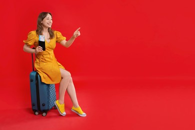 Photo of Happy traveller with passport and ticket sitting on suitcase against red background, space for text