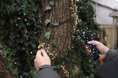 Photo of Man decorating tree trunk with Christmas lights outdoors, closeup