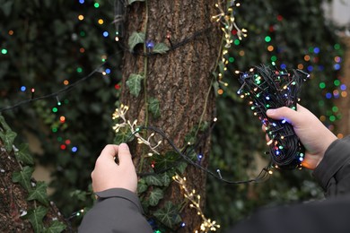 Photo of Man decorating tree trunk with Christmas lights outdoors, closeup