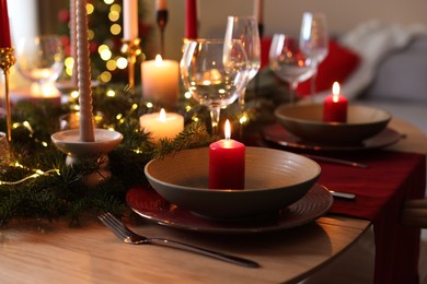 Photo of Beautiful dishware with burning candles, fir tree branches and Christmas lights on wooden table indoors, closeup