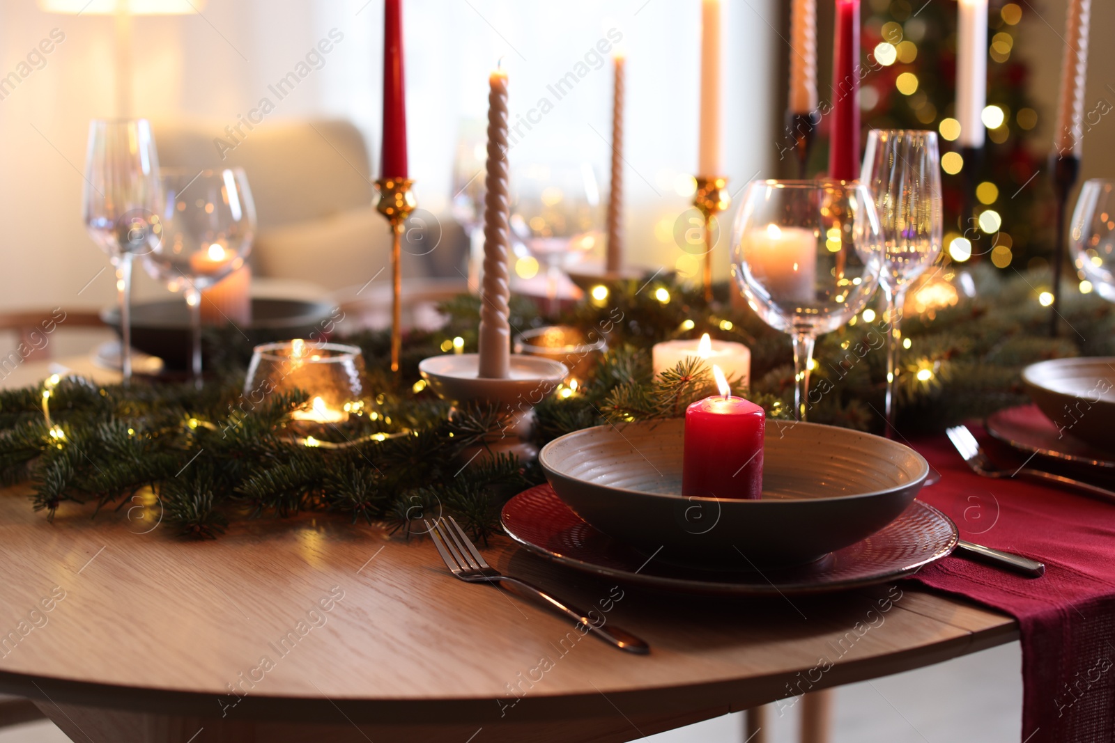 Photo of Beautiful dishware with burning candles, fir tree branches and Christmas lights on wooden table indoors, closeup