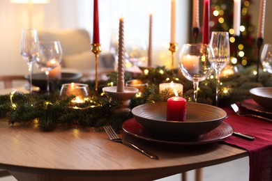 Photo of Beautiful dishware with burning candles, fir tree branches and Christmas lights on wooden table indoors, closeup