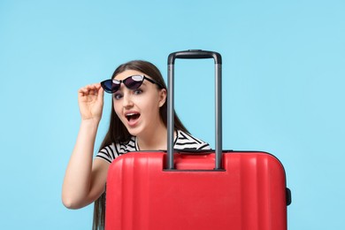 Emotional woman with suitcase wearing sunglasses on light blue background